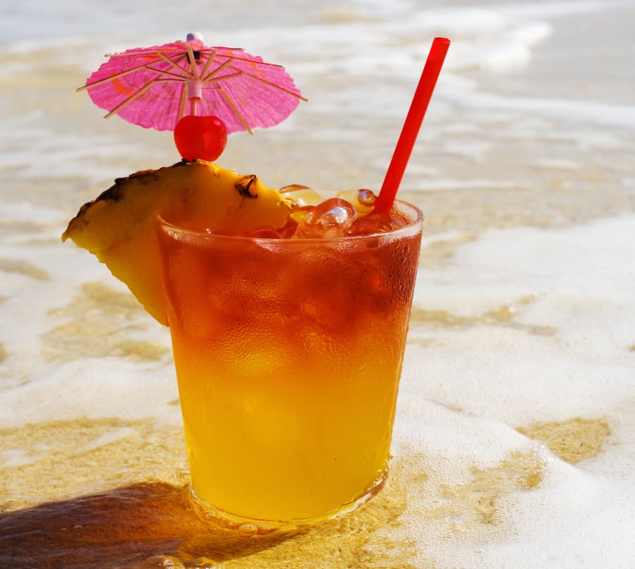 A mai tai garnished with pinapple and a cherry, sitting in shallow water on the beach.