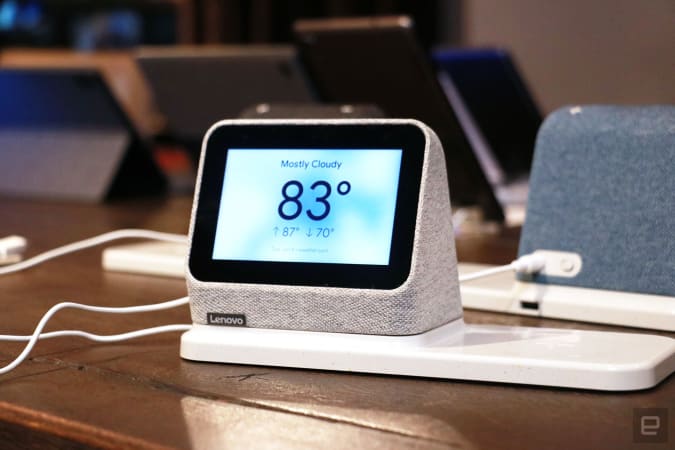 Off angle view of a gray Lenovo Smart Clock 2 on a wireless charging dock with its screen facing the camera. In the background sits a blue Smart Clock 2 and a row of tablets.
