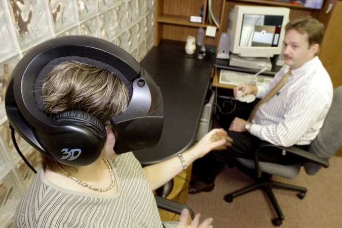 FOR USE WITH FEATURE TITLED VIRTUAL THERAPY -- Esther Begle, left, wears a virtual-reality helmet as she demonstrates a therapy session at Virtually Better Inc. in Atlanta, May 23, 2000, to combat her fear of flying. She reaches toward Ken Graap, CEO of Virtually Better, who uses a computer to control the simulated flight that Begle experiences.  (AP Photo/Ric Feld)