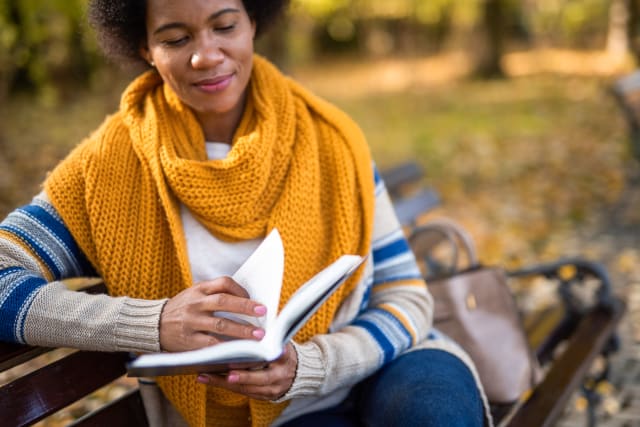 Mid adult African American woman sitting on bench and reading a book on autumn day in the park.