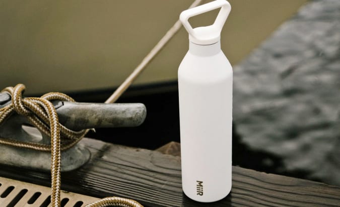 A white MiiR Insulated bottle sits on the edge of a dock with water and a boat in the background.