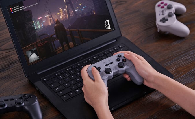 Two hands hold an 8BitDo Pro 2 game controller on a laptop keyboard, while a video game is displayed on the screen.