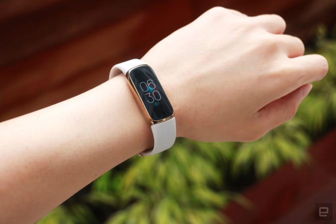 Slightly angled view of the Fitbit Luxe with a light pink silicone strap on a wrist against a dark brown background with some greenery.  The screen indicates that it is 6.30 p.m.
