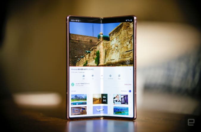 The Samsung Galaxy Z Fold 2 standing in portrait orientation on a wooden table with its screen partly folded and facing the camera. It shows a YouTube video playing. 