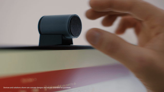 A barrel-shaped webcam sitting on a cradle on top of a monitor, with a hand reaching out to grab it. The camera is facing backwards.