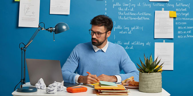Image of a man wearing glasses, a sweater and a button down shirt sitting at a desk with a pen in his hand.