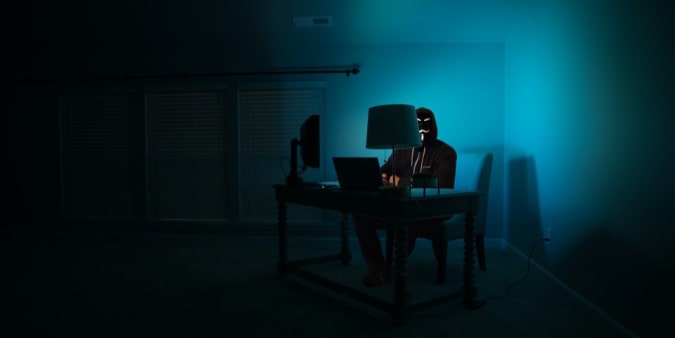 Stock image of a man in a mask at a computer, sitting in the dark.