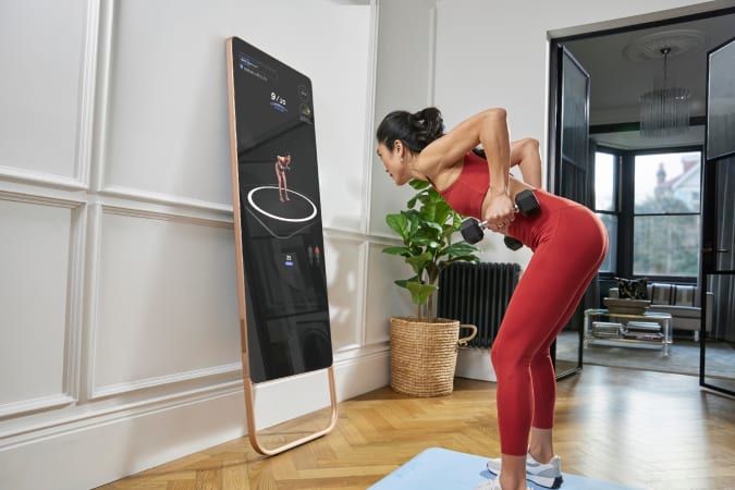 A woman holding dumbbells in the middle of a bent row, standing in front of a Fiture interactive mirror leaning against a wall. 