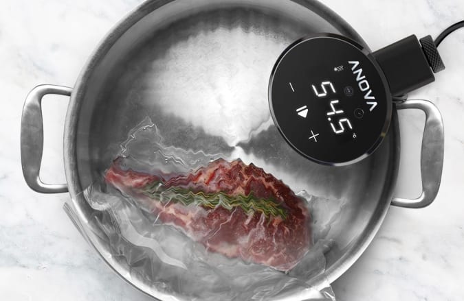 Anova Precision Cooker Nano sitter i en kastrull med vatten och tillagar en köttbit.