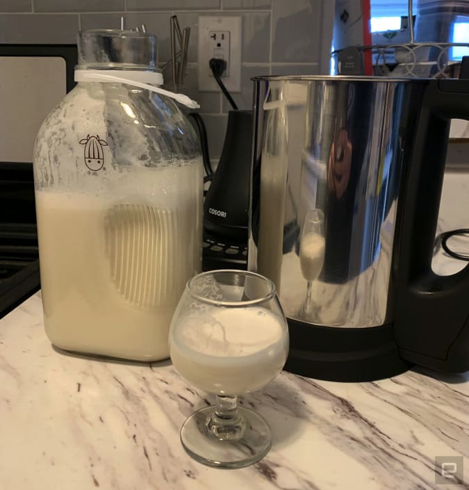 The Almond Cow plant milk maker next to a jug of homemade homemade cashew milk and a glass with cashew milk on a marbled countertop.