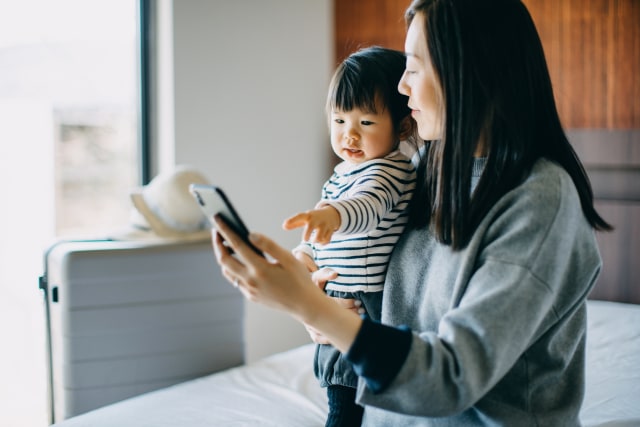 Young Asian mother and cute little daughter using smartphone in hotel room while on vacation