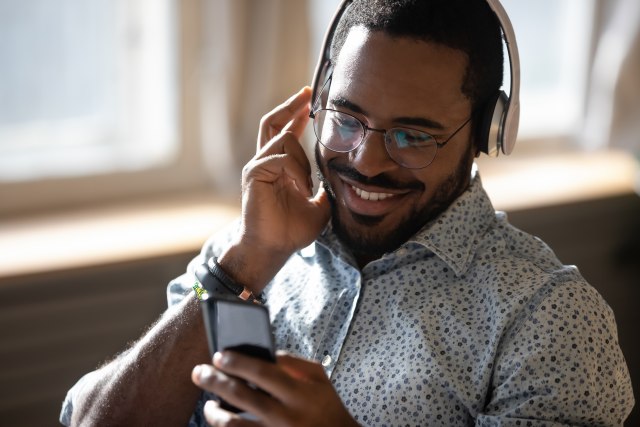 Smiling young adult african man wearing wireless headphones listening mobile music on smartphone, happy afro american guy using online player podcast audio book in app on cell phone relaxing at home