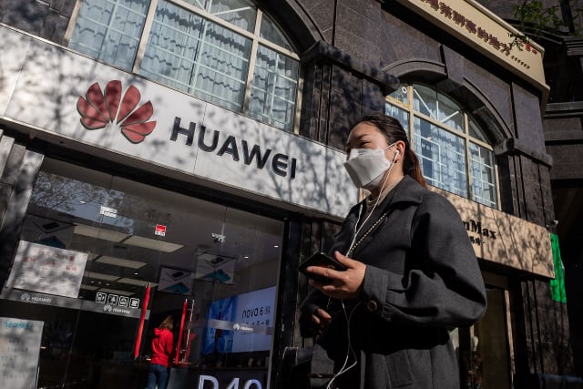 A woman wearing a face mask amid concerns over the COVID-19 coronavirus walks holding her smartphone past a Huawei shop (L) on a street in Beijing on April 22, 2020. - China's economy shrank for the first time in decades last quarter as the coronavirus paralysed the country, in a historic blow to the Communist Party's pledge of continued prosperity in return for unquestioned political power. (Photo by NICOLAS ASFOURI / AFP) (Photo by NICOLAS ASFOURI/AFP via Getty Images)