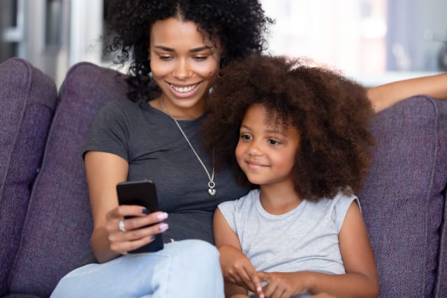 Happy afro mother or elder sister with little kid sitting on sofa in living room at home spend time using phone app having fun, watching cartoons make videocall, modern tech weekend activities concept